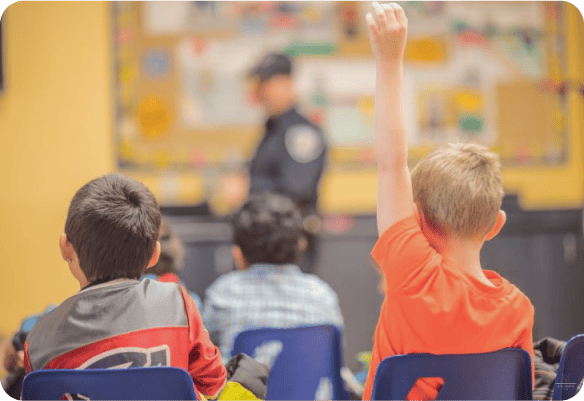 Children raising hands in classroom with police officer